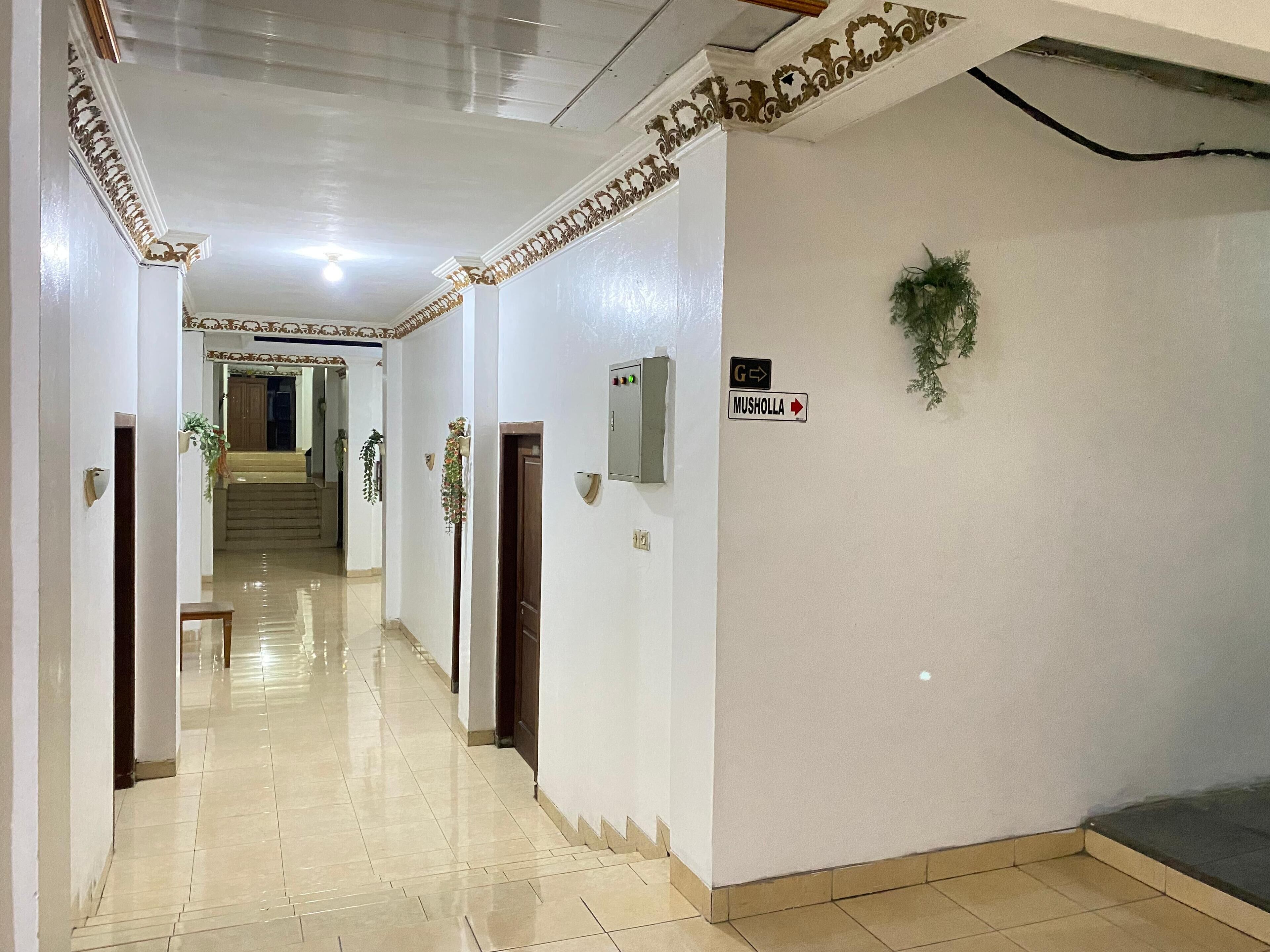 A hallway with white walls, a tiled floor, and a staircase at the end. There are signs indicating directions to 'G' and 'Musholla'.