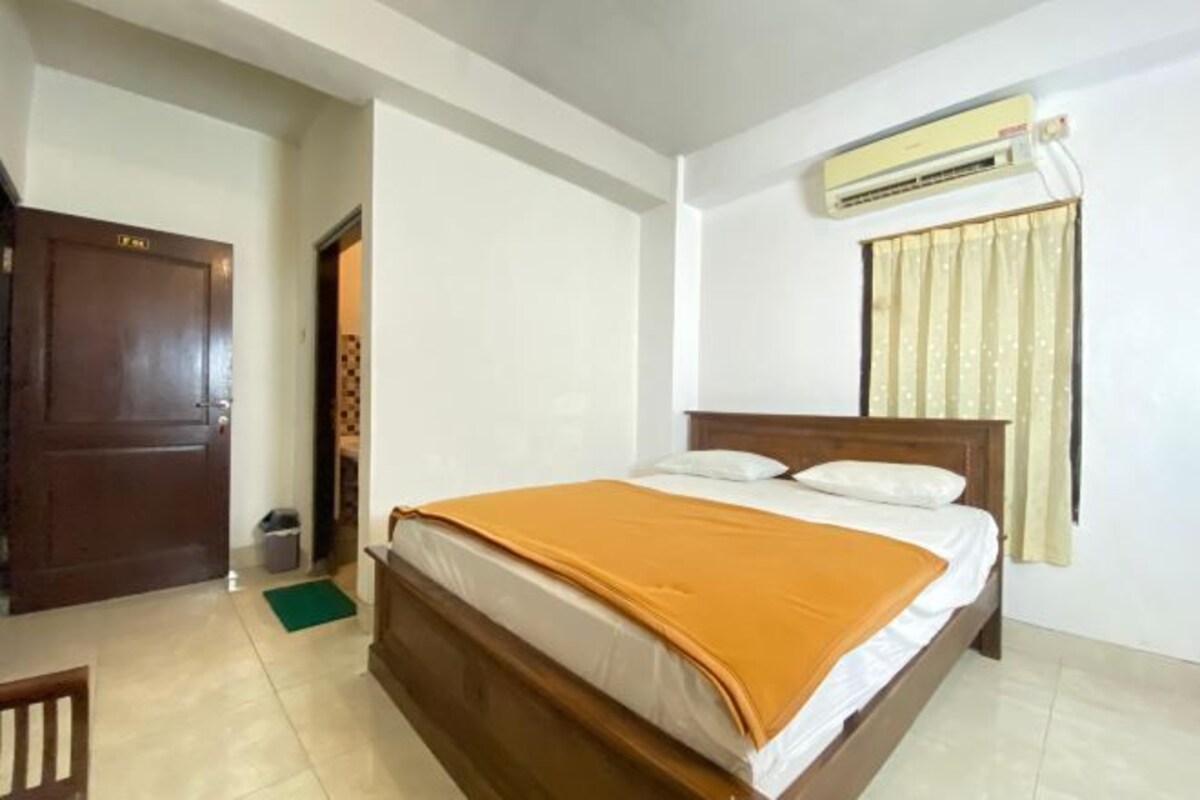 A bedroom with a wooden bed, a green mat, a white wall, a window with curtains, and an air conditioning unit.