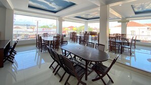 A spacious dining area with multiple tables and chairs, large windows offering views of the city, and a modern ceiling design. - New Wijaya Hotel (Kalasan)
