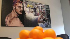 Et maleri af to muskuløse mænd i et fitnesscenter, med en skål appelsiner i forgrunden.