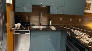 Une cuisine compacte avec des meubles de couleur turquoise, un lave-vaisselle en acier inoxydable et un plan de travail noir à motifs évoquant le marbre.