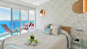A bedroom with a large bed, a view of the sea, a balcony with chairs, and a glass table with a tray of fruits and a plant.