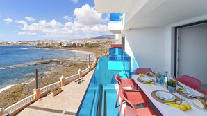 A balcony with a dining setup overlooking a coastal view, including a pool and beach.