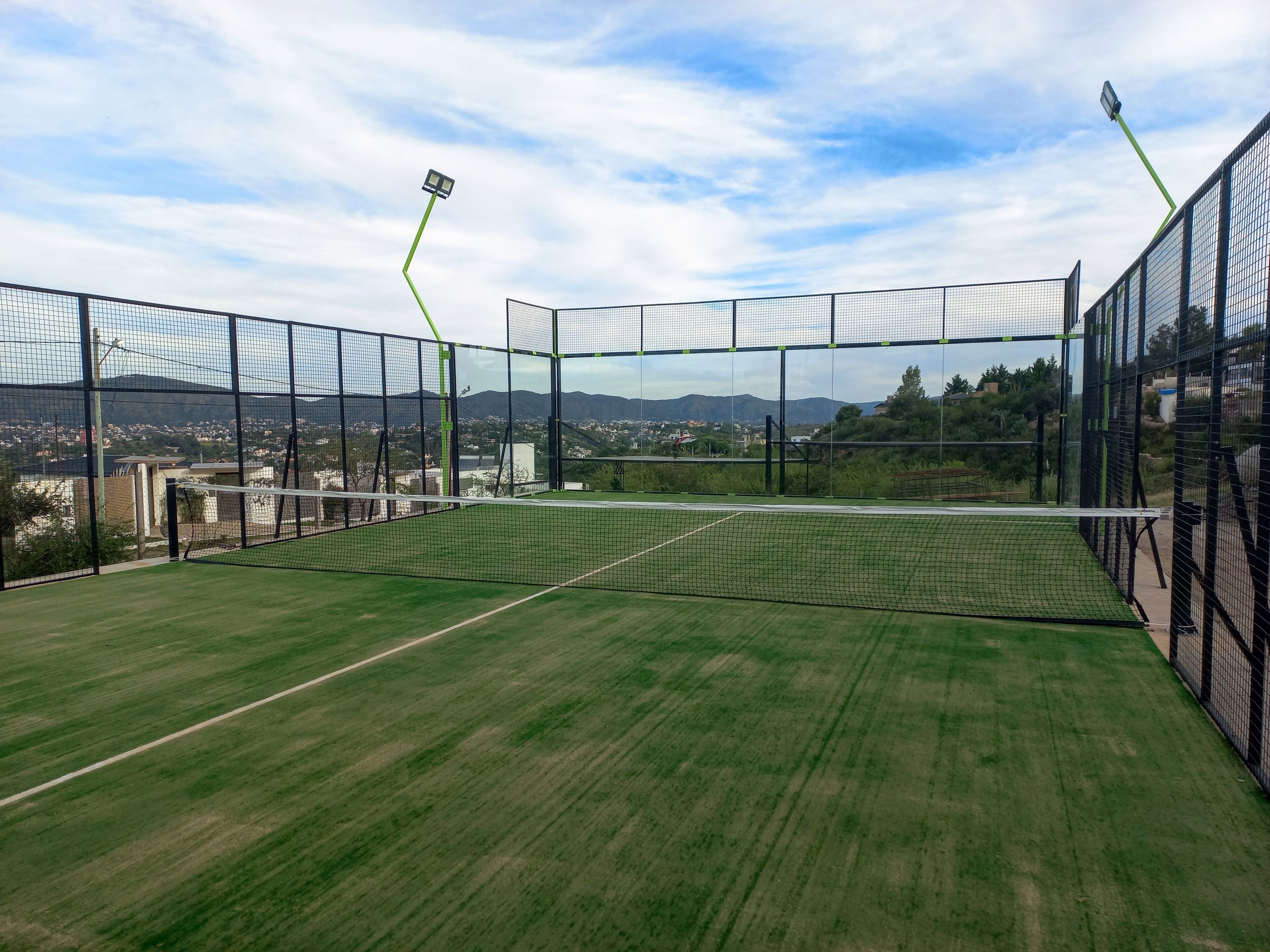 A tennis court with netted walls and floodlights, surrounded by a grassy field and a cityscape in the background.