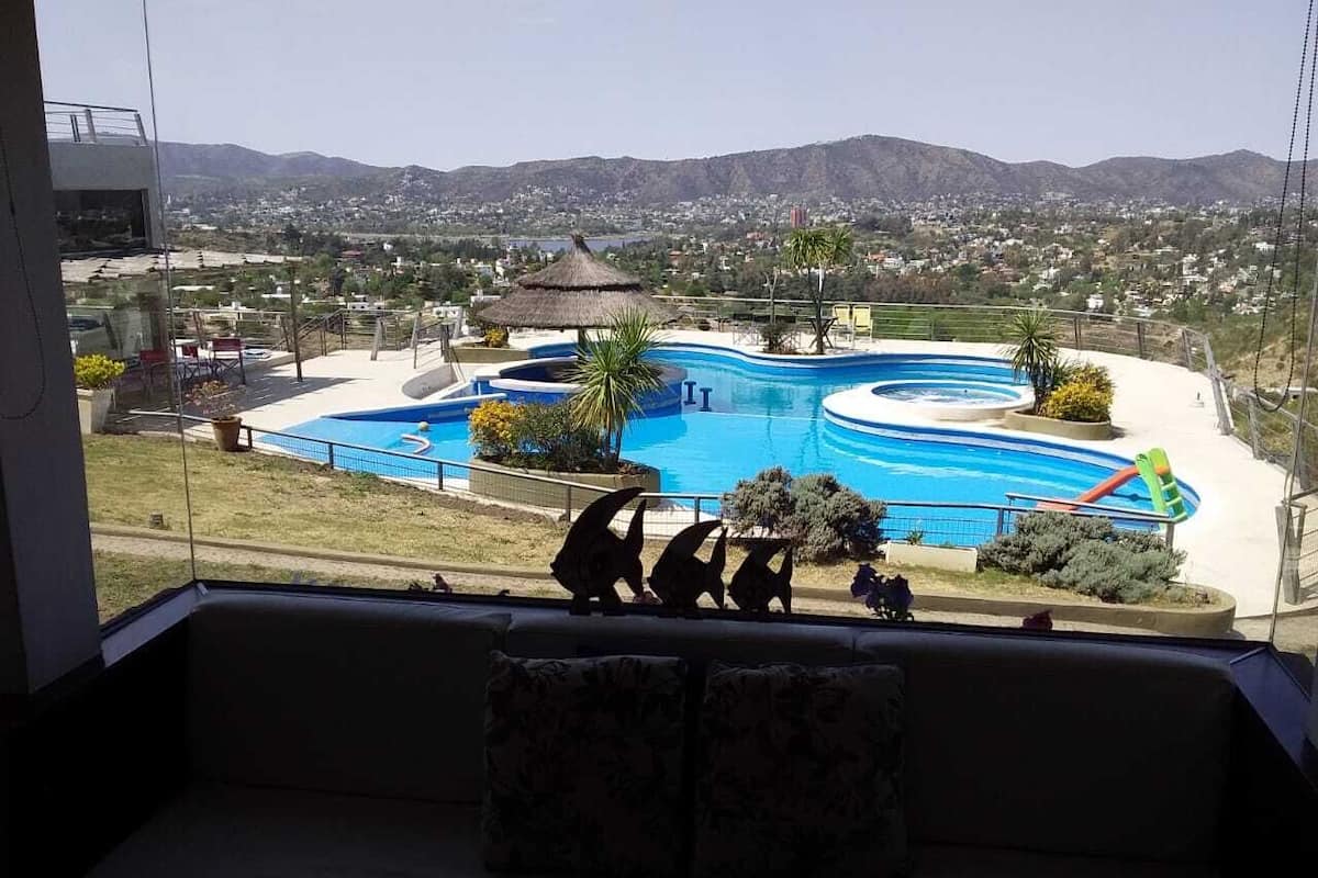 A view of a swimming pool with a slide, surrounded by greenery and a mountainous landscape.