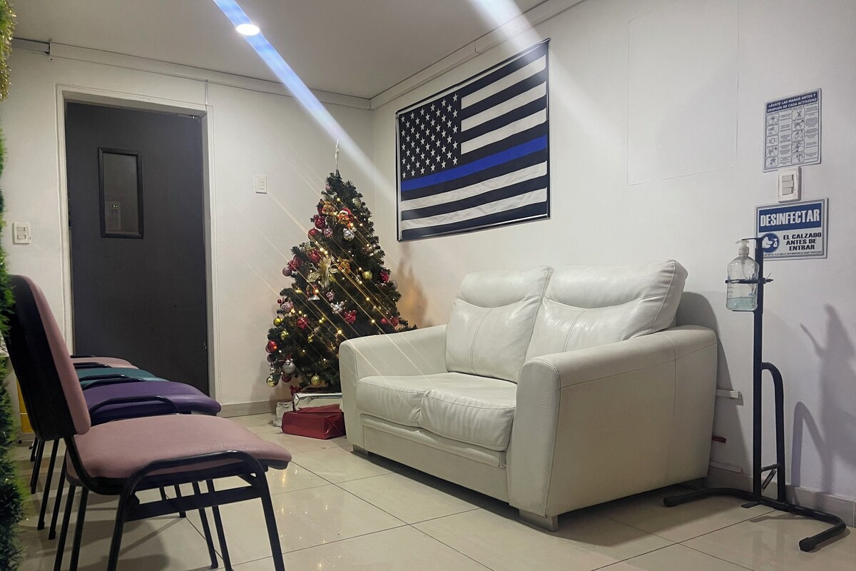 Une pièce avec un canapé en cuir blanc, un sapin de Noël, un drapeau accroché au mur et un distributeur de désinfectant.