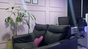 A modern living room with a black leather sofa, a large green plant, and a pink pillow. There are framed certificates on the wall and a black office chair in the background. - A 5 minutos del aeropuerto (Bogotá)