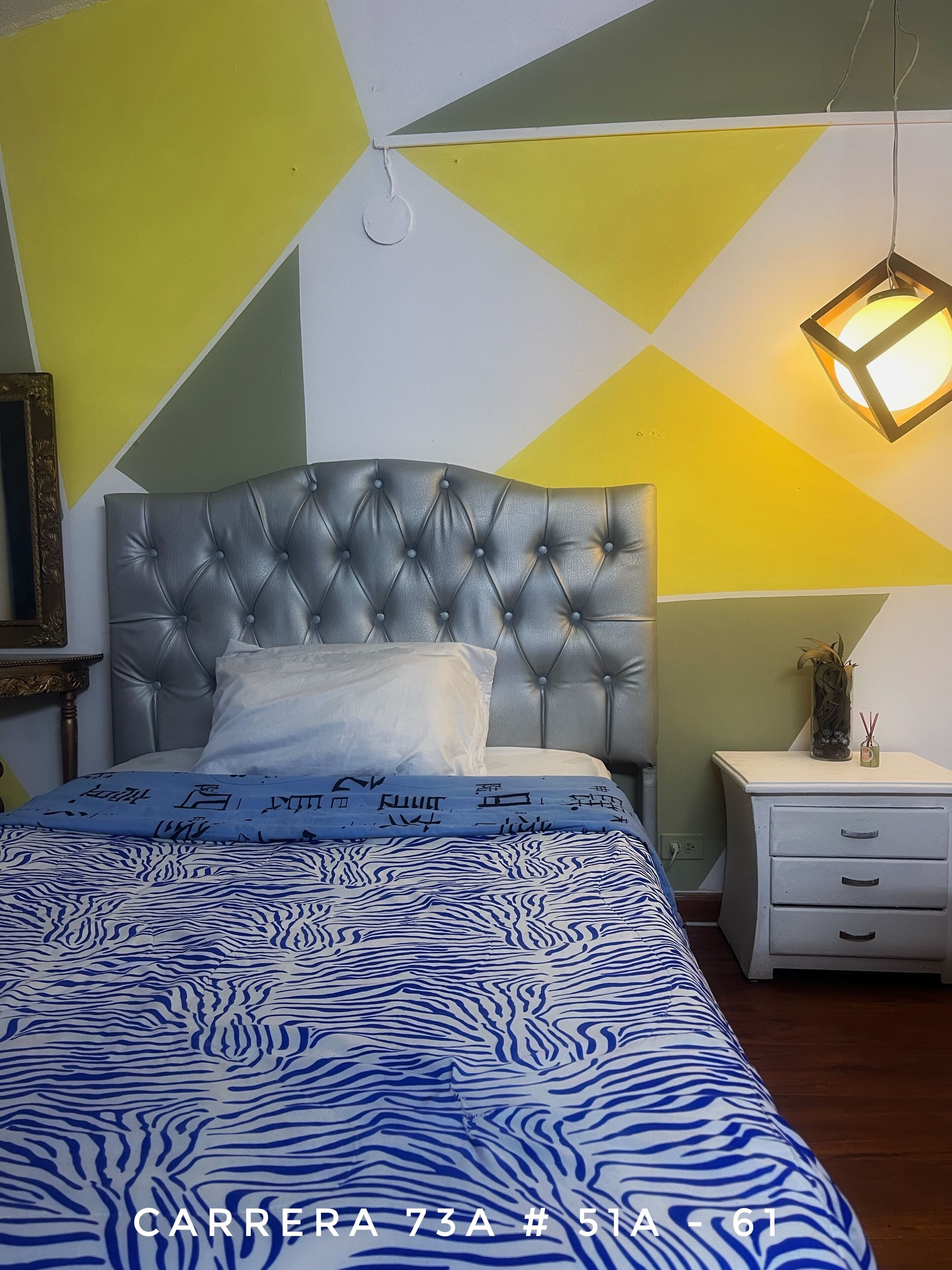 A bedroom with a tufted headboard, a patterned bedspread, a bedside table with a vase, and a geometric wall design.