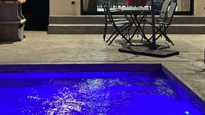 A swimming pool with blue lighting, a patio chair, a table, and a view of a dining area through a window. - Villas Ambiance 21 Elégance Méditerranéenne à Portiragnes, Entre Terre et mer (Portiragnes)