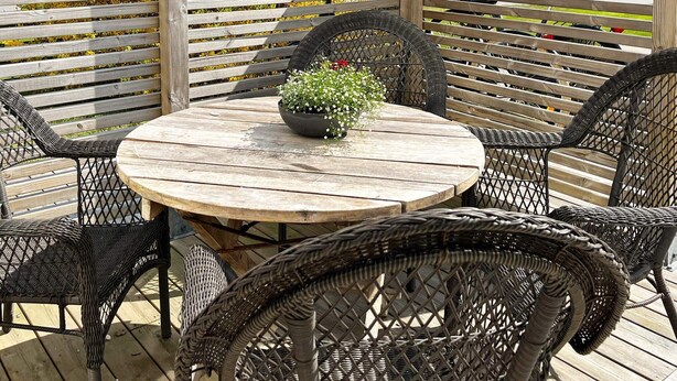 A round wooden table with a black wicker chair around it, set on a wooden deck with a wooden fence in the background. - 4 Person Holiday Home in Kungshamn (Kungshamn)