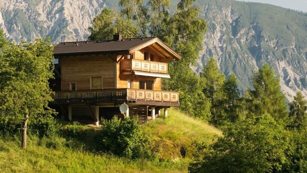 Une cabane en bois à deux étages, dotée d’un balcon, entourée d’une végétation luxuriante et avec des montagnes en toile de fond.
