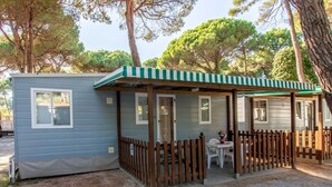 A small, single-story mobile home with a striped awning and a fenced patio area. - Rivaverde Family Camping Village Lodge Deluxe für 5 Personen (Marina di Ravenna)
