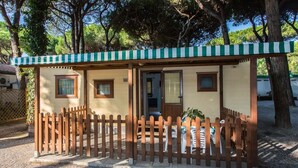 A small, single-story structure with a green and white striped awning, a wooden fence, and a door. - Rivaverde Family Camping Village Lodge Superior für 3 Personen (Marina di Ravenna)