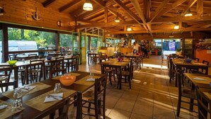 A rustic wooden interior with multiple dining tables and chairs, large windows, and a high ceiling with exposed beams. - Rivaverde Family Camping Village Lodge Superior für 3 Personen (Marina di Ravenna)