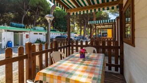 A covered outdoor seating area with a checkered tablecloth, wooden chairs, and a wooden fence. - Rivaverde Family Camping Village Lodge Superior für 3 Personen (Marina di Ravenna)