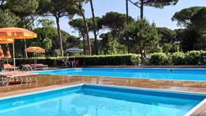 A rectangular swimming pool with a wooden deck, surrounded by lounge chairs and umbrellas. - Rivaverde Family Camping Village Lodge Superior für 3 Personen (Marina di Ravenna)