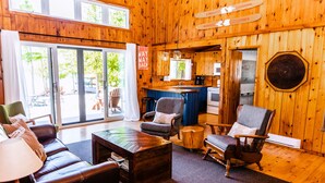 Ein gemütliches Blockhaus-Interieur mit Wohnbereich, inklusive Sofa, Sesseln und Couchtisch. Eine Küchenzeile mit Spüle und Herd ist sichtbar, und ein großes Fenster bietet Blick ins Freie.