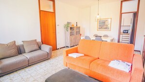 Een woonkamer met een grijze bank, een oranje bank, een houten kast en een eethoek met een tafel en stoelen.