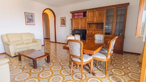 Uma sala de jantar com mesa e cadeiras de madeira, um sofá, um armário com portas de vidro e ladrilhos de chão estampados.
