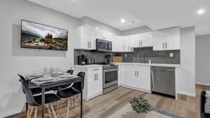 A modern kitchen with white cabinets, a stove, microwave, and dishwasher. There is a dining table with chairs and a potted plant on the floor. - ModernSuite*NearAirport*2 Queens*ROKU (Edmonton)