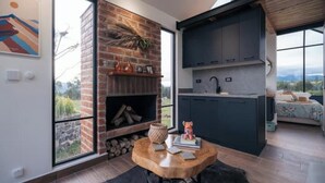 A modern kitchen with dark cabinets and a brick fireplace, a round wooden table with books and a decorative item, and a large window offering a view of the outdoors. - House with mountain views

 (Checa)