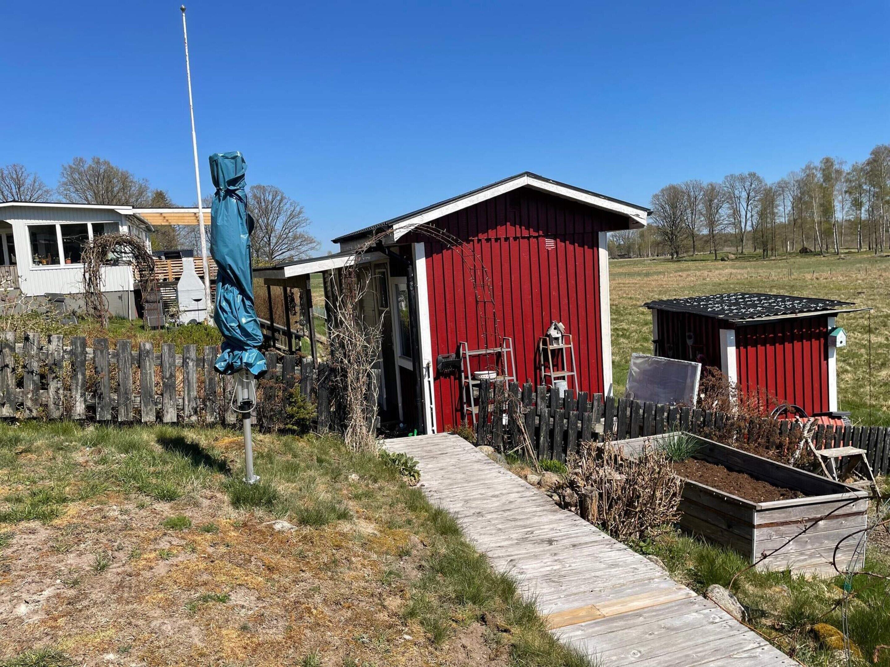 A red wooden shed with a door, a small attached structure, a wooden fence, and a wooden pathway leading to the shed.
