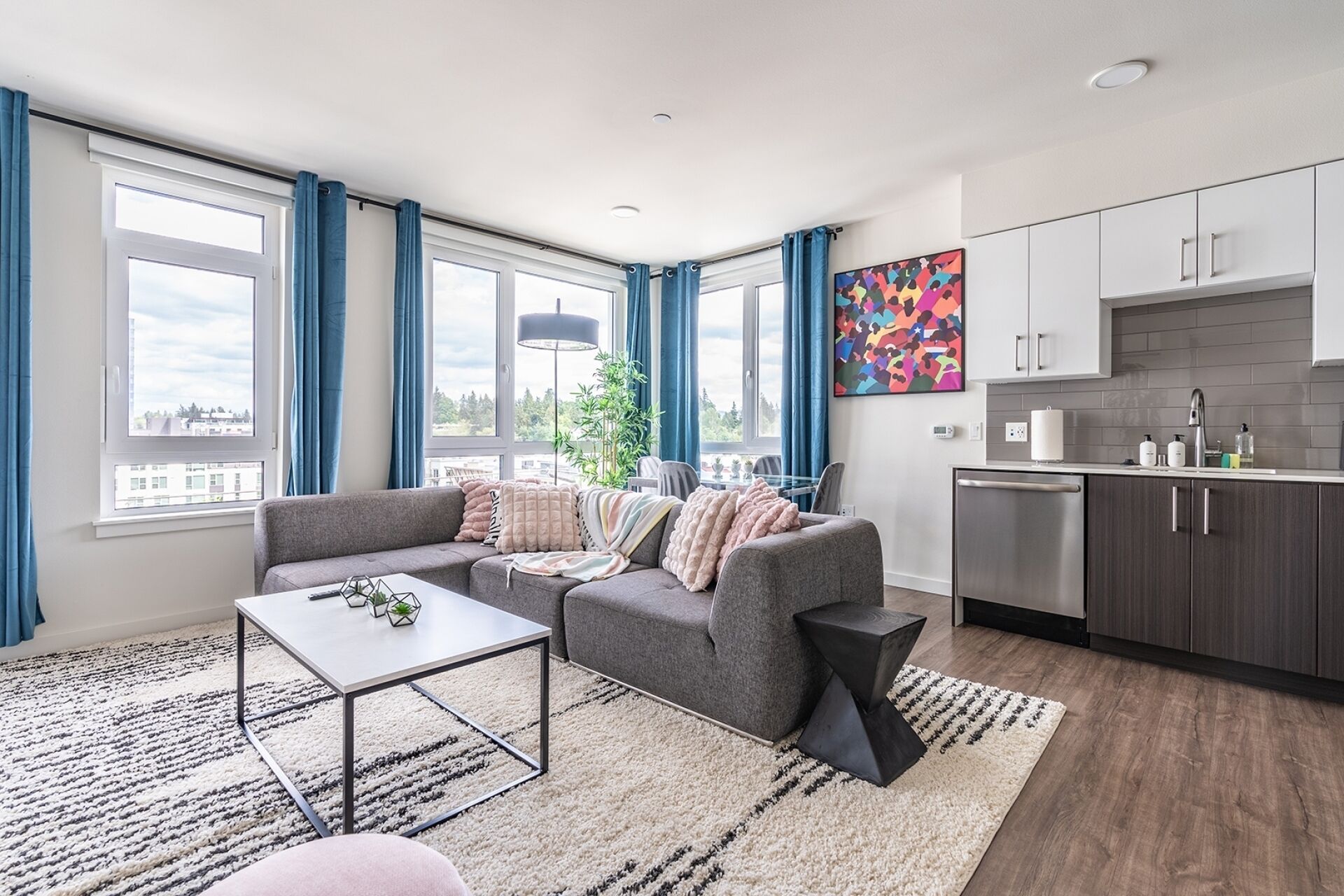 En modern lägenhet med en grå soffa, ett matbord, ett kök med grå skåp och stora fönster med blå gardiner.