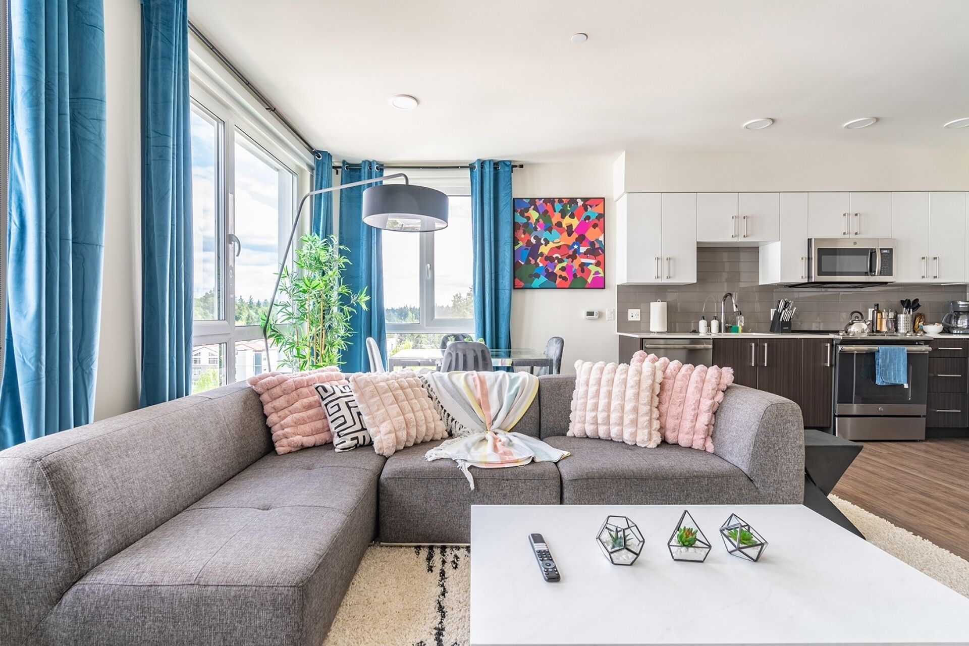 A modern living room with a grey sectional sofa, a white coffee table, and a kitchen area with white cabinets and a microwave.