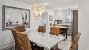 A dining area with a glass table and wicker chairs, a chandelier, and a maritime painting on the wall.
