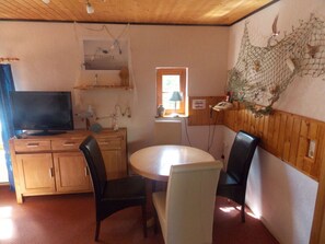 A room with wooden cabinets, a television, a round table with chairs, and a window with a view of greenery. - Strandfleh Modern Retreat (Wangerooge)