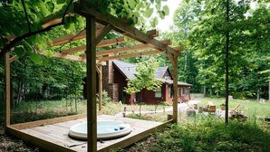 Une terrasse en bois avec un jacuzzi, entourée d’une pergola en bois, située dans un espace boisé avec un chalet en arrière-plan.