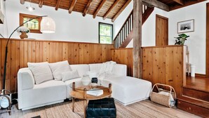 Un salon chaleureux avec un canapé d’angle blanc, une table basse ronde en bois et un pouf noir. On y trouve également un panier en osier et un lampadaire.
