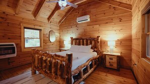 Un dormitorio de cabaña de madera con una cama, dos mesitas de noche, un ventanal, un equipo de aire acondicionado y un ventilador de techo.