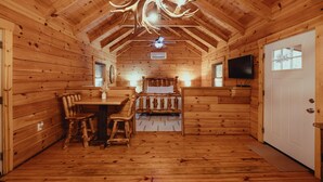 Interior de una cabaña de troncos con comedor, cama, ventilador de techo y televisión.