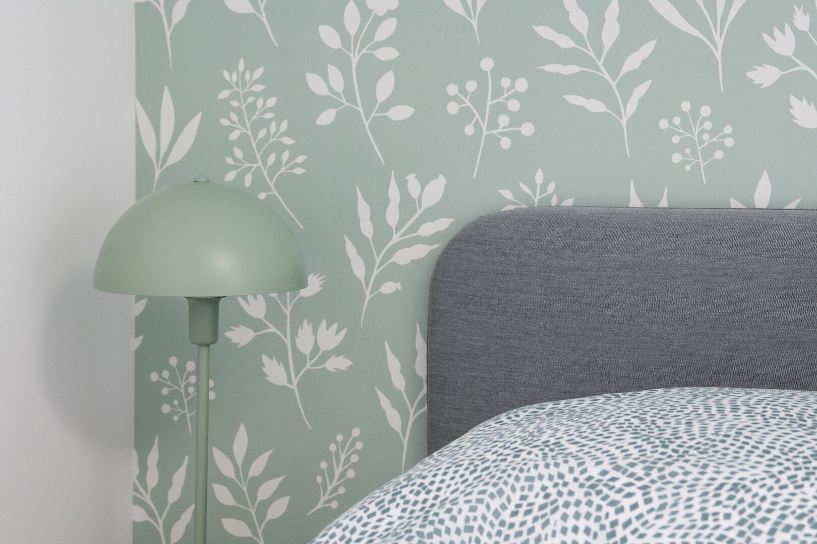A room with a green patterned wallpaper, a grey headboard, and a patterned bedspread.