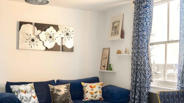 Un salon moderne avec un canapé bleu, des coussins décoratifs, une table basse en verre et une grande fenêtre ornée de rideaux à motifs.