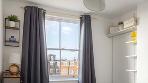 Une chambre à coucher avec un lit, une fenêtre avec des rideaux, une étagère avec une plante et une lampe.