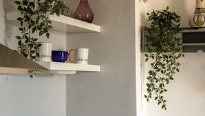 Une cuisine avec des murs recouverts de carreaux blancs, un robinet en acier inoxydable et des étagères où sont rangés divers objets, notamment des tasses et un vase.