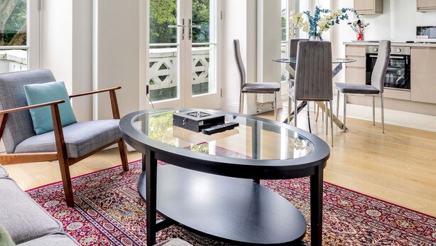 A modern living room with a glass coffee table, grey upholstered chairs, and a red patterned rug. - Balcony South Kensington Luxury 2 bed Apartment (London)