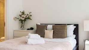 A neatly made bed with a pink blanket, a bedside table with a vase of flowers, and a nightstand with a phone. - Balcony South Kensington Luxury 2 bed Apartment (London)