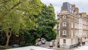 A view of a street with parked cars and a building with a brick facade and multiple windows. - Balcony South Kensington Luxury 2 bed Apartment (London)