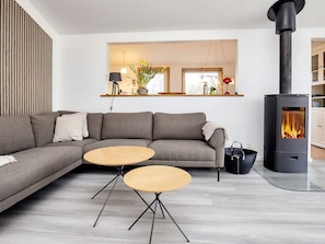 A modern living room with a sectional sofa, two round wooden tables, a black wood-burning stove, and a large window with a view of the outdoors. - 4 Star Holiday Home in Blokhus (Blokhus)