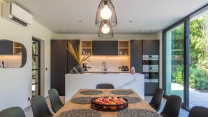 A modern kitchen with a central island, a dining table with placemats and a fruit bowl, and a large window offering a view of greenery. - Luxury Villa With Pool & Sauna Kortrijk (Kortrijk)