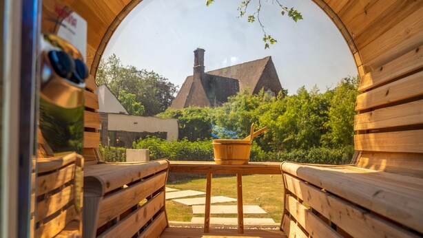 A wooden sauna with benches and a table, a wooden bench with a pair of sunglasses, and a view of a house and greenery outside. - Luxury Villa With Pool & Sauna Kortrijk (Kortrijk)