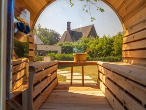 A wooden sauna with benches and a table, a wooden bench with a pair of sunglasses, and a view of a house and greenery outside. - Luxury Villa With Pool & Sauna Kortrijk (Kortrijk)