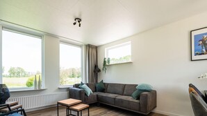 A modern living room with a sectional sofa, a small wooden table, and two windows offering a view of greenery. - Cozy Holiday Home in Oploo Near Forest (Oploo)