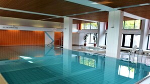 Une piscine intérieure à l’eau d’un bleu cristallin, entourée de murs blancs et de poutres en bois au plafond.