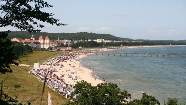 A beach with a long stretch of umbrellas and sun loungers, a pier extending into the water, and buildings in the background. - Sandpiper in Haus Strandburg Binz (Binz)