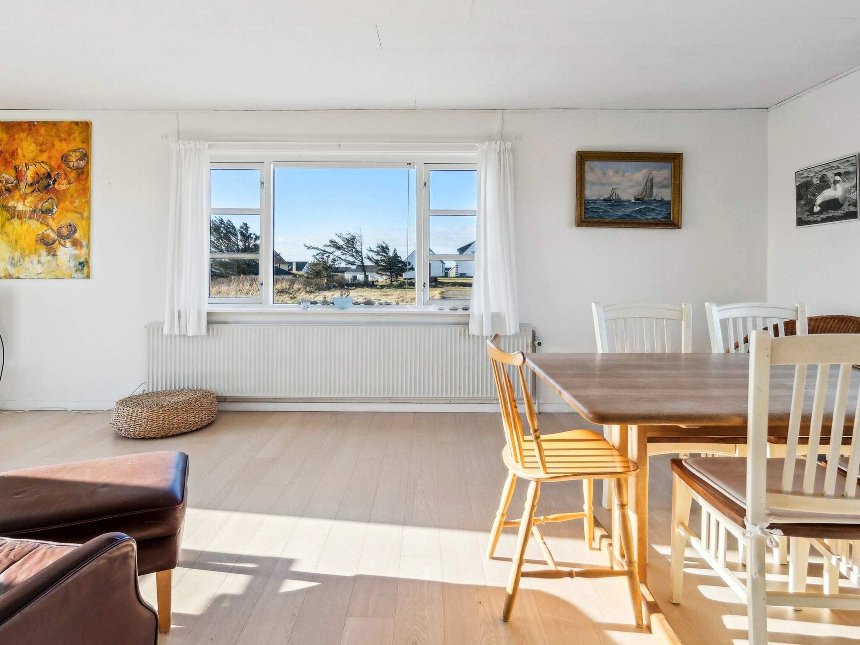 A dining area with a wooden table and chairs, a large window with a view of the outdoors, and artwork on the walls.