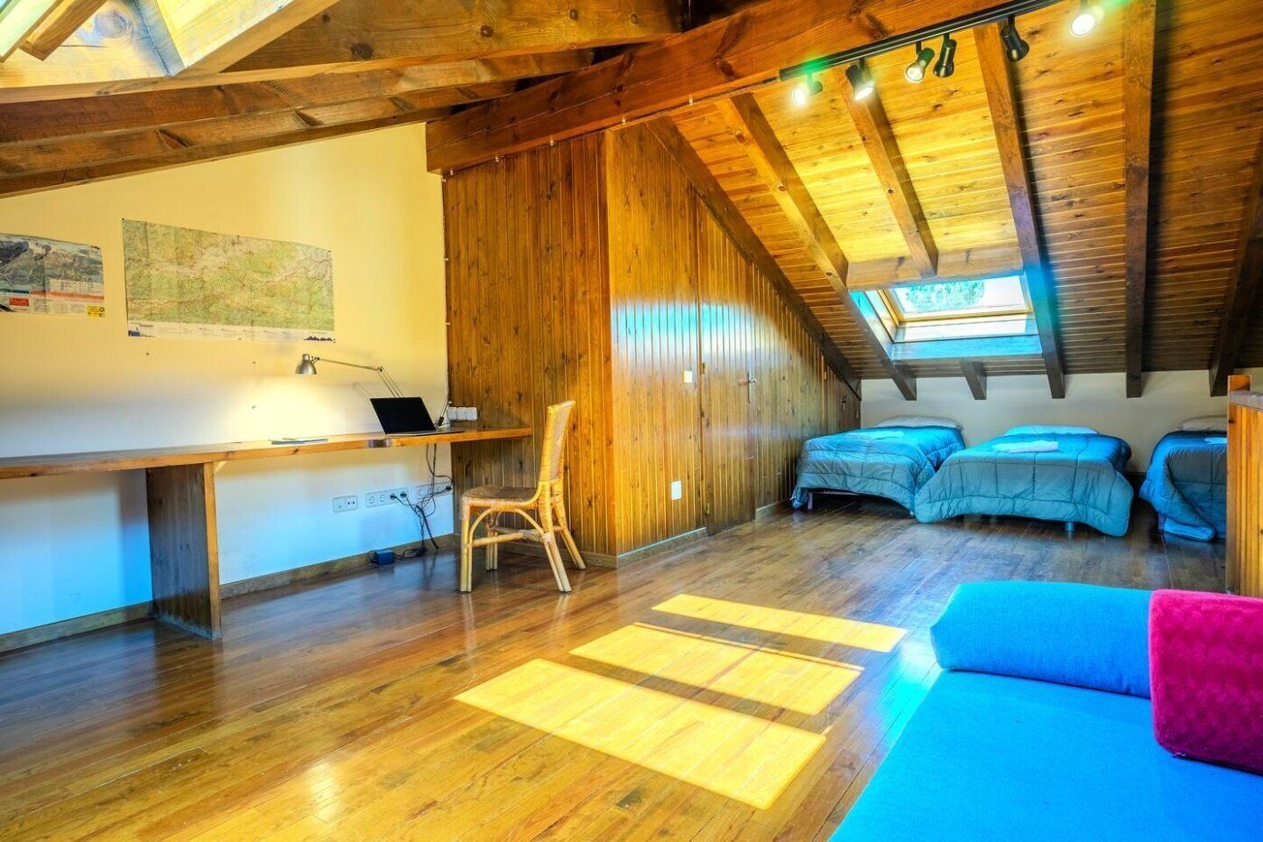Quarto de sótão acolhedor, com vigas de madeira, uma escrivaninha com um laptop, uma cadeira e três camas.
